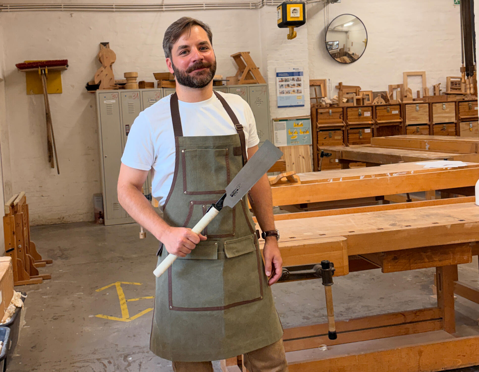 Roma in the woodworking workshop Roma is standing in the woodworking workshop wearing a white shirt and an olive green apron. in his right hand he is holding a Japanese saw.
